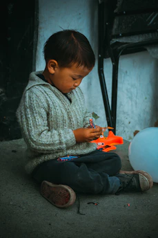 A young child sits cross-legged on the ground, engrossed in playing with a small, colorful toy. The child wears a gray knitted sweater and dark pants, with slightly worn shoes. A bright orange toy lies nearby, and a white balloon rests on the ground next to the child. The background shows a textured wall and a folded chair.