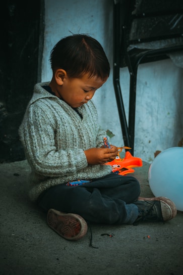 A young child sits cross-legged on the ground, engrossed in playing with a small, colorful toy. The child wears a gray knitted sweater and dark pants, with slightly worn shoes. A bright orange toy lies nearby, and a white balloon rests on the ground next to the child. The background shows a textured wall and a folded chair.
