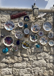 A stone wall adorned with decorative plates featuring various artistic designs, including images of fruits, sea creatures, and abstract patterns. A hanging lantern is present, along with a ceramic seagull and a wooden sign. There are also some small ceramic items with vibrant colors.