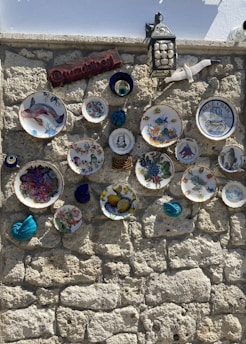 A stone wall adorned with decorative plates featuring various artistic designs, including images of fruits, sea creatures, and abstract patterns. A hanging lantern is present, along with a ceramic seagull and a wooden sign. There are also some small ceramic items with vibrant colors.