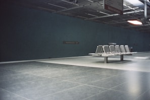 A deserted subway or train station platform with metal benches and a tiled floor. The space is dimly lit with a dark green wall and a sign indicating the station name. Above, a partially visible ceiling structure and signage add to the minimalist, utilitarian design.
