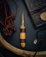 An intricately crafted hunting knife with a rustic wooden handle and a somewhat tarnished blade is at the center. It is surrounded by a leather-bound book with embossed designs, a leather pouch tied with cords, a carved antler, and a small metallic pendant with an engraved symbol. The composition exudes a vintage and adventurous aesthetic.