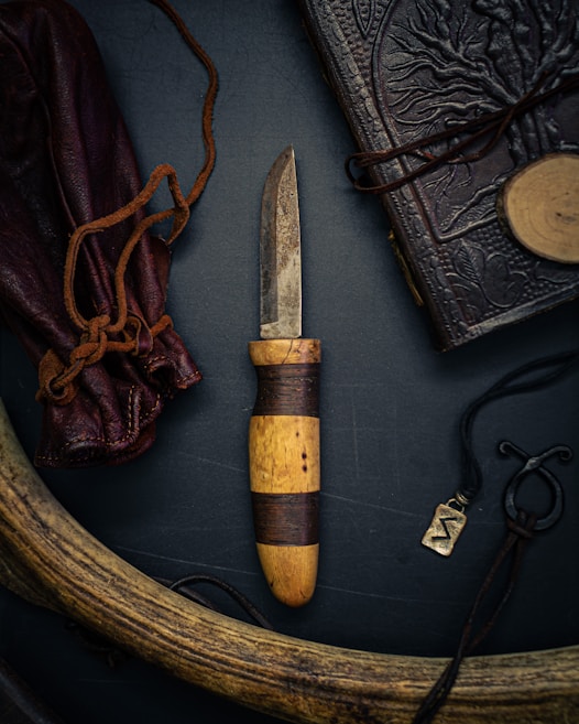 An intricately crafted hunting knife with a rustic wooden handle and a somewhat tarnished blade is at the center. It is surrounded by a leather-bound book with embossed designs, a leather pouch tied with cords, a carved antler, and a small metallic pendant with an engraved symbol. The composition exudes a vintage and adventurous aesthetic.