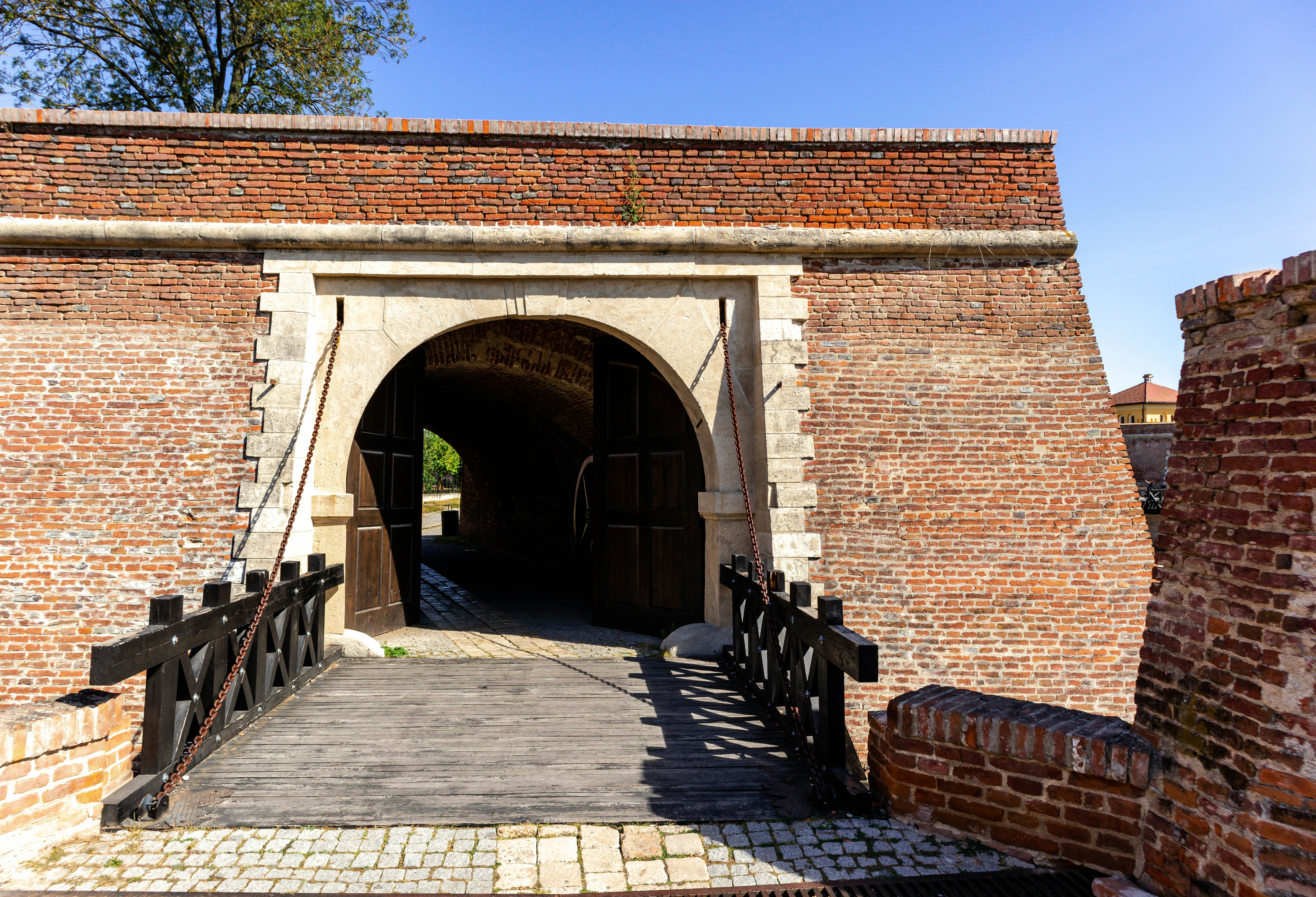 Brick arch gate with a wooden ramp leading to a shadowed passage, framed by sunlit brick and stone under a clear blue sky.
