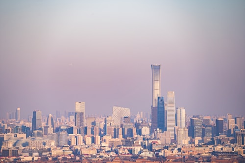 A panoramic cityscape featuring a skyline with numerous high-rise buildings and skyscrapers. The tallest building is a modern, sleek tower dominating the view, surrounded by other structures of varying architectural styles. The scene is set under a clear sky with a subtle gradient transitioning from blue to pink.