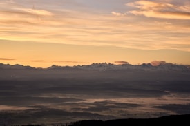 A panoramic landscape at sunset showcases a serene view of distant mountain ranges under a sky painted with soft, warm hues of orange and yellow. The foreground features rolling hills partially veiled in mist, creating a tranquil and dreamy atmosphere.