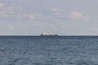A large cargo ship is seen on the horizon of a vast, calm ocean. The sky above is filled with scattered clouds, and the water reflects a subtle blue-gray hue.