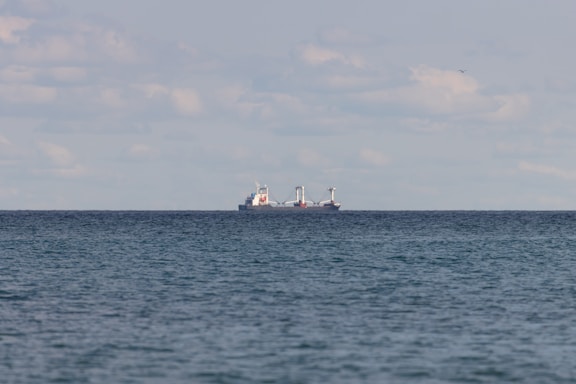 A large cargo ship is seen on the horizon of a vast, calm ocean. The sky above is filled with scattered clouds, and the water reflects a subtle blue-gray hue.