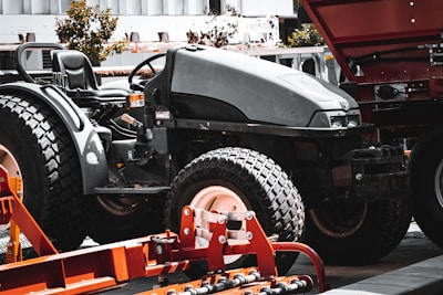A large black tractor with big, textured tires is parked in an industrial outdoor setting. Adjacent to the tractor, there is a piece of orange agricultural machinery with metal components. In the background, parts of a building and some trees are visible.