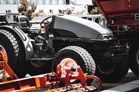A large black tractor with big, textured tires is parked in an industrial outdoor setting. Adjacent to the tractor, there is a piece of orange agricultural machinery with metal components. In the background, parts of a building and some trees are visible.