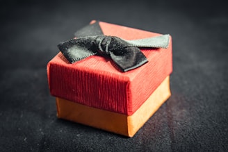 A small, elegant gift box with a red lid and a golden base is adorned with a black bow. The texture of the paper is slightly ribbed, adding a tactile element. The box sits against a dark, blurred background.