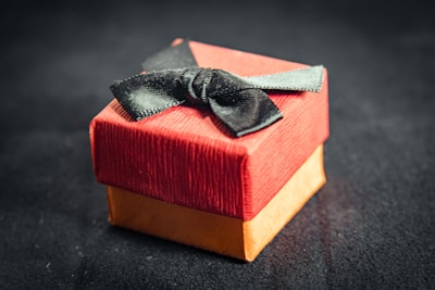 A small, elegant gift box with a red lid and a golden base is adorned with a black bow. The texture of the paper is slightly ribbed, adding a tactile element. The box sits against a dark, blurred background.