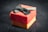 A small, elegant gift box with a red lid and a golden base is adorned with a black bow. The texture of the paper is slightly ribbed, adding a tactile element. The box sits against a dark, blurred background.