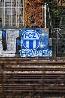 Graffiti art on a concrete wall features a blue and white FCZ logo above the German words 'F&uuml;r Immer'. The wall is located alongside train tracks, with a partially dried climbing plant in the background. Metal fencing and some urban greenery surround the scene, contributing to an urban and gritty atmosphere.