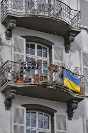 An ornate building facade with arched windows and decorative wrought iron balconies. A Ukrainian flag is draped over the railing of one balcony, which is also adorned with potted plants and miscellaneous objects.
