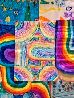 A collection of vibrant, multicolored drawings featuring abstract patterns and shapes. The central piece showcases dynamic, curved lines with a rainbow spectrum, surrounded by other artworks with similar vivid hues and whimsical designs.