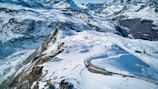 A scenic mountainous landscape featuring snow-covered peaks and a winding red train moving along a railway track. Skiers or hikers are visible on the slopes, complementing the vastness of the rugged terrain. The backdrop includes clear blue skies and distant mountain ranges.