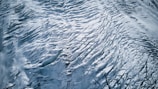 An aerial view of a glacier with intricate patterns and textures. The surface features crevasses and ridges formed by the movement of ice over time. The monochromatic palette emphasizes the stark beauty and vastness of the icy landscape.