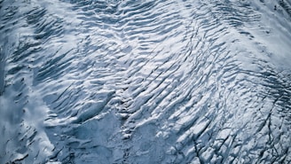An aerial view of a glacier with intricate patterns and textures. The surface features crevasses and ridges formed by the movement of ice over time. The monochromatic palette emphasizes the stark beauty and vastness of the icy landscape.