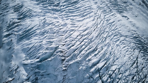 An aerial view of a glacier with intricate patterns and textures. The surface features crevasses and ridges formed by the movement of ice over time. The monochromatic palette emphasizes the stark beauty and vastness of the icy landscape.
