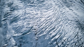 An aerial view of a glacier with intricate patterns and textures. The surface features crevasses and ridges formed by the movement of ice over time. The monochromatic palette emphasizes the stark beauty and vastness of the icy landscape.