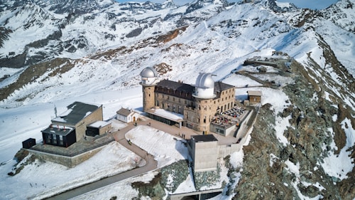 A large observatory building with two domes located on a snowy mountain peak. Surrounding the structure are snowy slopes and rocky outcrops, providing a panoramic view of the high-altitude landscape. Several smaller buildings are nearby, and a terrace with tables and chairs is visible on one side of the observatory, indicating a place for visitors.