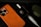 An orange smartphone case with an Apple logo, a white stylus, a winged skull brooch in a black box, and an olive green rugged case are placed on a dark surface.