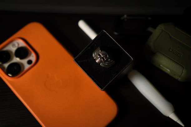 An orange smartphone case with an Apple logo, a white stylus, a winged skull brooch in a black box, and an olive green rugged case are placed on a dark surface.