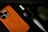 An orange smartphone case with an Apple logo, a white stylus, a winged skull brooch in a black box, and an olive green rugged case are placed on a dark surface.