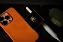 An orange smartphone case with an Apple logo, a white stylus, a winged skull brooch in a black box, and an olive green rugged case are placed on a dark surface.