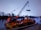 A large crane mounted on a floating barge is lifting equipment on a calm river. The scene is set against a backdrop of trees and a distant bridge, with a soft, overcast sky.