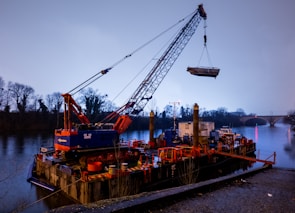 A large crane mounted on a floating barge is lifting equipment on a calm river. The scene is set against a backdrop of trees and a distant bridge, with a soft, overcast sky.
