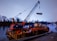 A large crane mounted on a floating barge is lifting equipment on a calm river. The scene is set against a backdrop of trees and a distant bridge, with a soft, overcast sky.