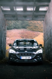 A car with a visible license plate is parked under a roofed passageway surrounded by stone walls. In the background, there is a mix of grass, shrubs, and autumn-colored foliage.