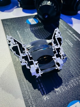 A cross-section of a camera lens is placed on a dark surface with charts and numbers, highlighting the internal structure and lens elements. The background seems to contain other camera equipment, suggesting a technical or professional setting.