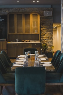 An elegant dining area featuring a wooden table set for eight, complete with glassware and plates. The table is surrounded by luxurious dark green chairs. The background includes a wooden cabinet with glass doors and a stone wall, enhancing the rustic and cozy ambiance.