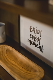 A framed motivational quote on a shelf reads 'enjoy every moment.' Next to it, there is a wooden tray with a light brown finish and a simple white mug placed on the shelf.