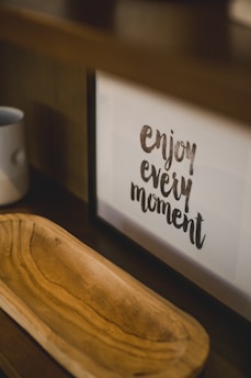 A framed motivational quote on a shelf reads 'enjoy every moment.' Next to it, there is a wooden tray with a light brown finish and a simple white mug placed on the shelf.