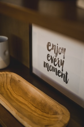 A framed motivational quote on a shelf reads 'enjoy every moment.' Next to it, there is a wooden tray with a light brown finish and a simple white mug placed on the shelf.