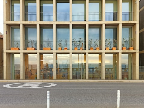 A modern building with large glass windows and a grid-like facade, featuring rows of planter boxes with plants. The building has a reflective surface, displaying a view of the horizon and sky. In front of the building, a street is visible with road markings and a traffic sign.