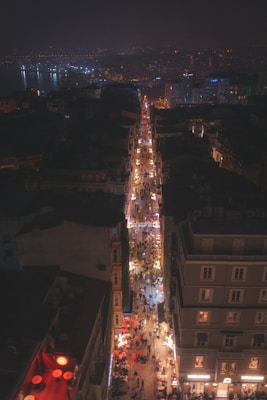 A bustling city street is illuminated by bright lights and filled with people, extending into the distance. Tall buildings frame the street, with warm tones and outdoor seating visible. City lights sparkle in the background, hinting at a vibrant nightlife.