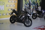 A sleek black Kawasaki motorcycle is prominently displayed indoors. The showroom features a bright yellow wall with the logo 'GM MOTO' repeated across it. In the background, another motorcycle can be observed along with a printed outdoor scene, suggesting an adventurous theme.