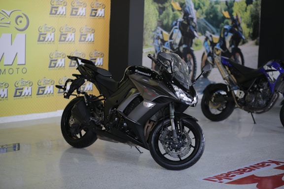 A sleek black Kawasaki motorcycle is prominently displayed indoors. The showroom features a bright yellow wall with the logo 'GM MOTO' repeated across it. In the background, another motorcycle can be observed along with a printed outdoor scene, suggesting an adventurous theme.