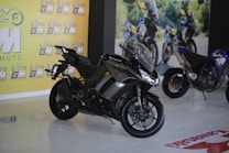 A sleek black Kawasaki motorcycle is prominently displayed indoors. The showroom features a bright yellow wall with the logo 'GM MOTO' repeated across it. In the background, another motorcycle can be observed along with a printed outdoor scene, suggesting an adventurous theme.