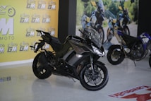 A sleek black Kawasaki motorcycle is prominently displayed indoors. The showroom features a bright yellow wall with the logo 'GM MOTO' repeated across it. In the background, another motorcycle can be observed along with a printed outdoor scene, suggesting an adventurous theme.