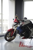 A sleek motorcycle with a red, white, and blue color scheme is positioned indoors near large windows. The motorcycle features prominent gold forks and black tires with a patterned tread. Natural light floods the space, highlighting the details of the motorcycle.