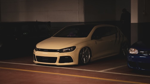 A white modified car with large wheels is parked in a dimly lit garage next to other vehicles. The car has a sleek design with a lowered body, and the setting is predominantly dark with artificial lighting highlighting the car's features.