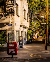 A quiet urban street scene with a pale building casting shadows from overhead wires and greenery. A red postal box with graffiti is prominent in the foreground, marked with a postal code. The street is lined with trees casting dappled sunlight and a few people are walking in the distance.