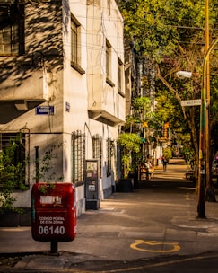 A quiet urban street scene with a pale building casting shadows from overhead wires and greenery. A red postal box with graffiti is prominent in the foreground, marked with a postal code. The street is lined with trees casting dappled sunlight and a few people are walking in the distance.