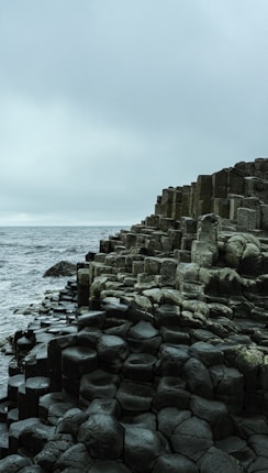 Hexagonal basalt columns on the edge of a rugged coastline meet a cloudy sky and choppy sea, creating a dramatic natural landscape.